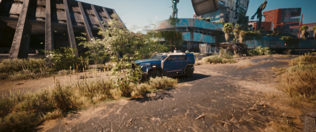 Cyberpunk 2077 screenshot: a blue SUV hides in the bushes next to some dilapidated buildings. 